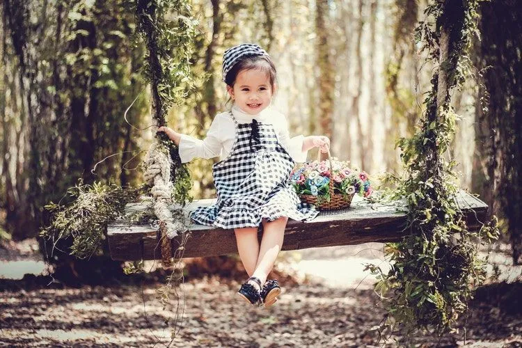 A little girl wearing checkered dress is sitting on a swing with flower basket