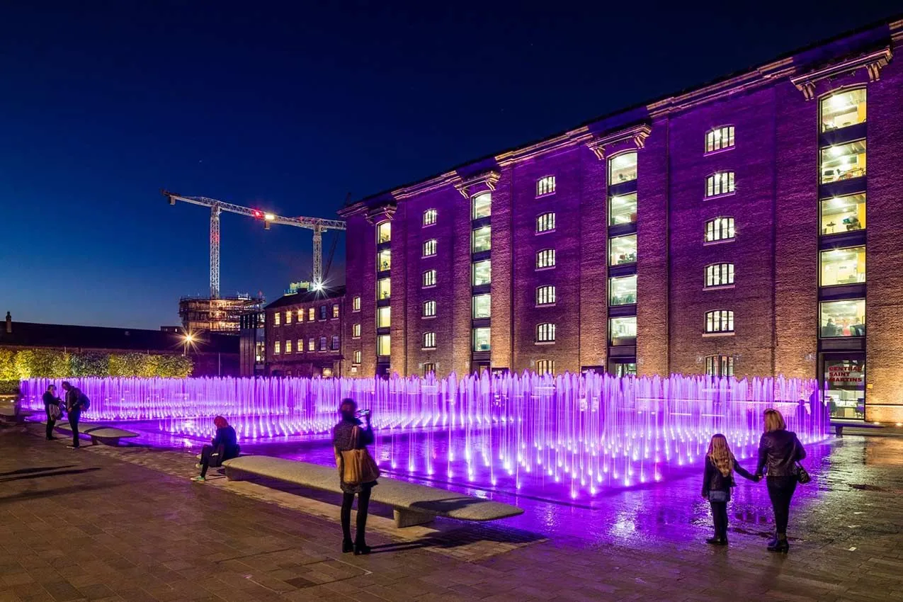 People looking at Granary Square fountains at night, lit up with purple LEDs