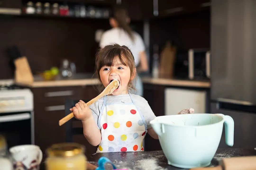A young girl licking the spoon after learning how to bake a delicious cake.