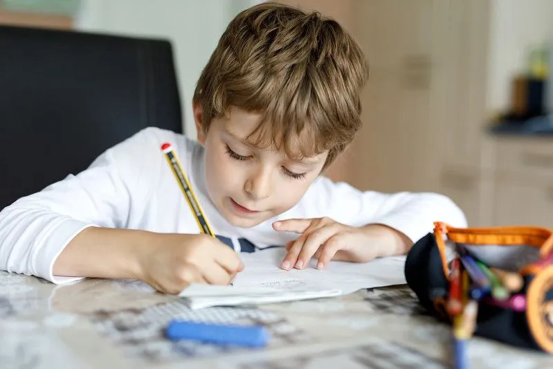 Young boy with light brown hair in white shirt sat at a desk, writing something in pencil.