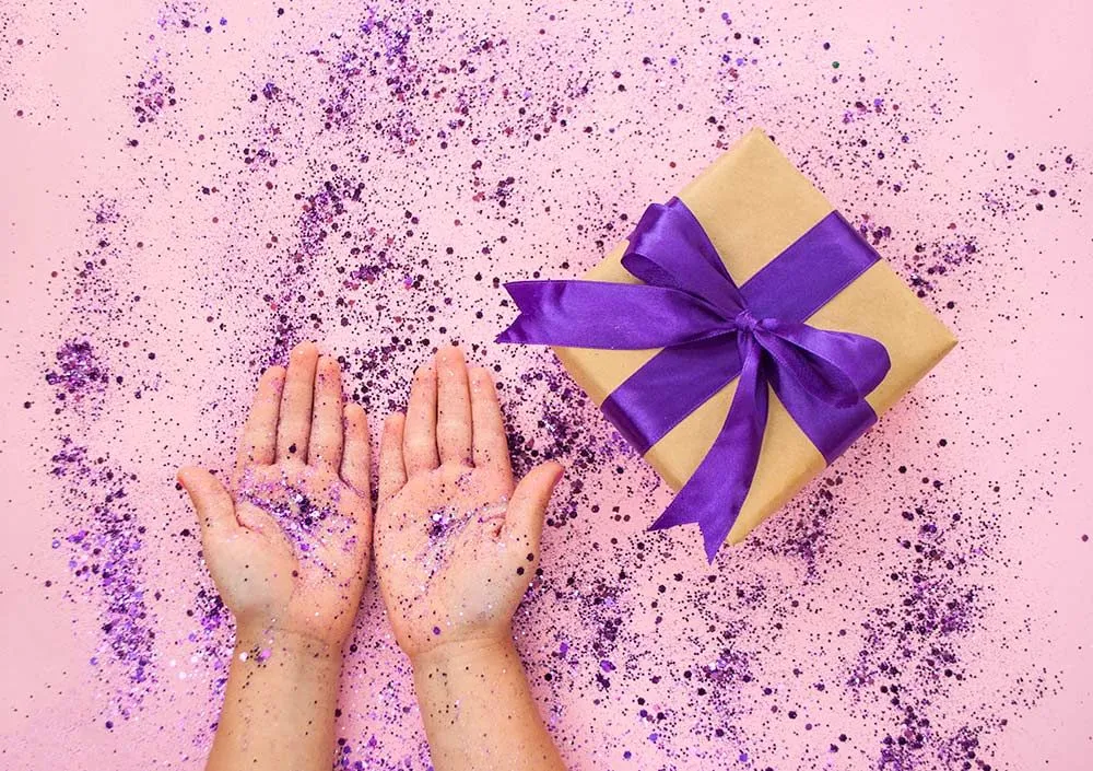 pink background with wrapped shoe box and hands covered in purple glitter
