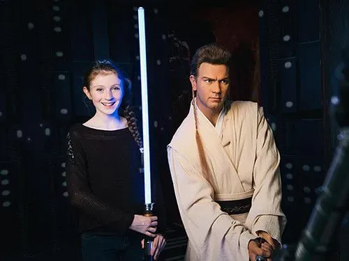 Girl standing next to Obi Wan Kenobi waxwork figure, holding a lightsaber.