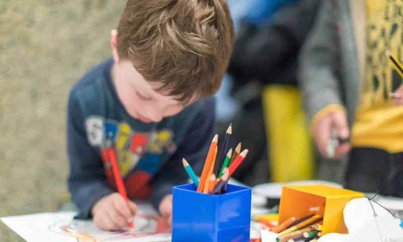 Young boy concentrating very hard on his drawing. A box of colourful pencils is in the foreground.