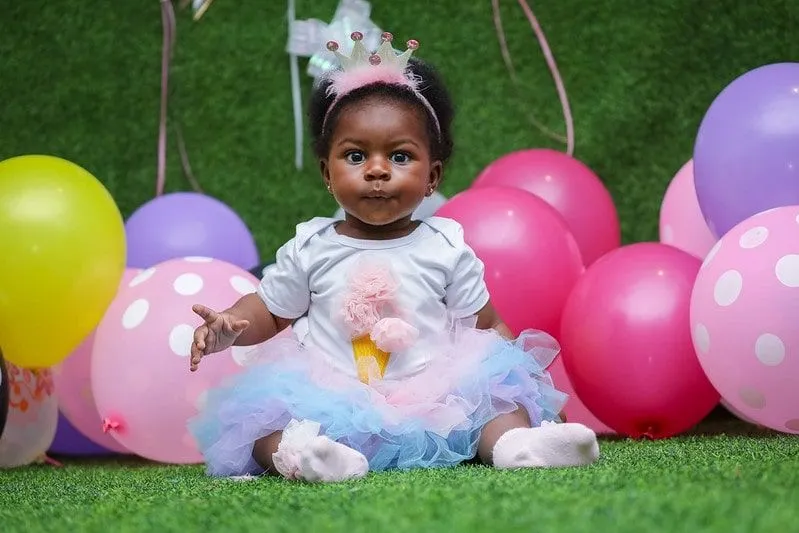 Baby girl wearing a hairband with a crown on it sat on the floor surrounded by balloons.