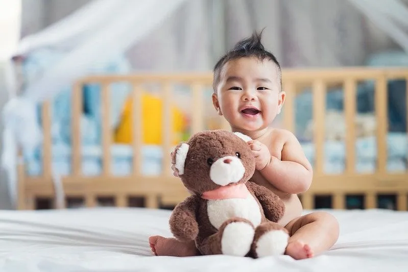 Baby boy sitting on the bed smiling with a teddy bear in arms.