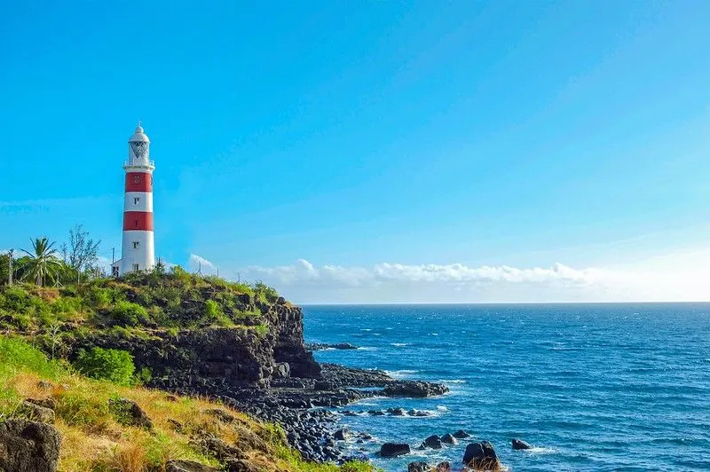 Red and white striped lighthouse on cliff, with bright blue sea to the left. It's a clear, sunny day and the sky is bright blue.