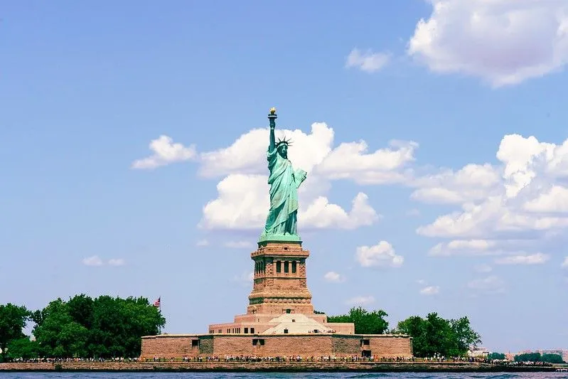 The Statue of Liberty in New York, USA, viewed from the front across the water. There is a blue sky with a few clouds in the background.