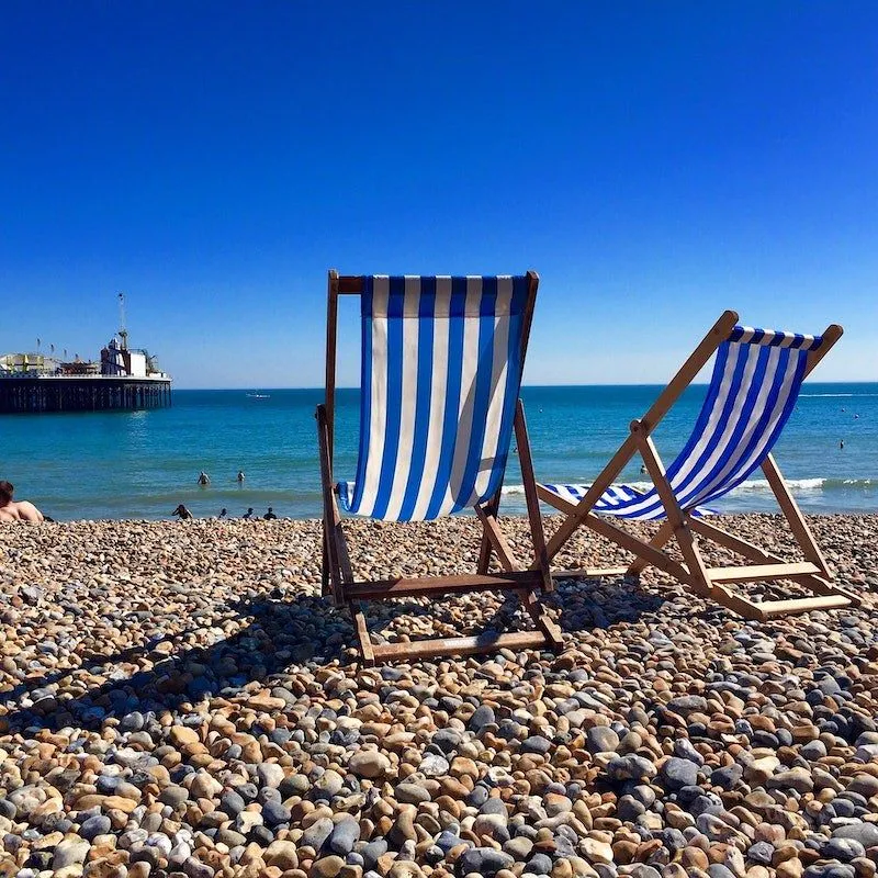 Two blue and white striped deck chairs on the pebbles of the beach, facing the sea on a sunny day.