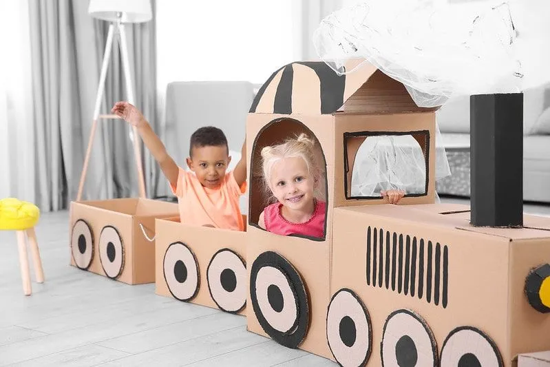 Children playing and smiling inside a cardboard train at home. 