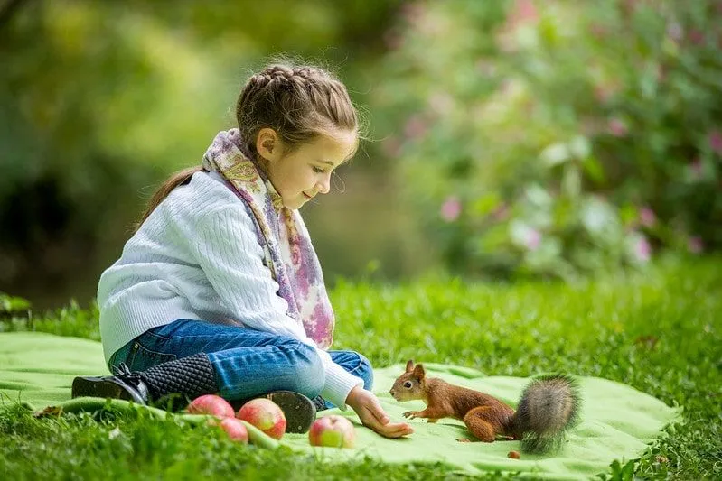 Little girl sitting on a rug on the grass feeding a squirrel.