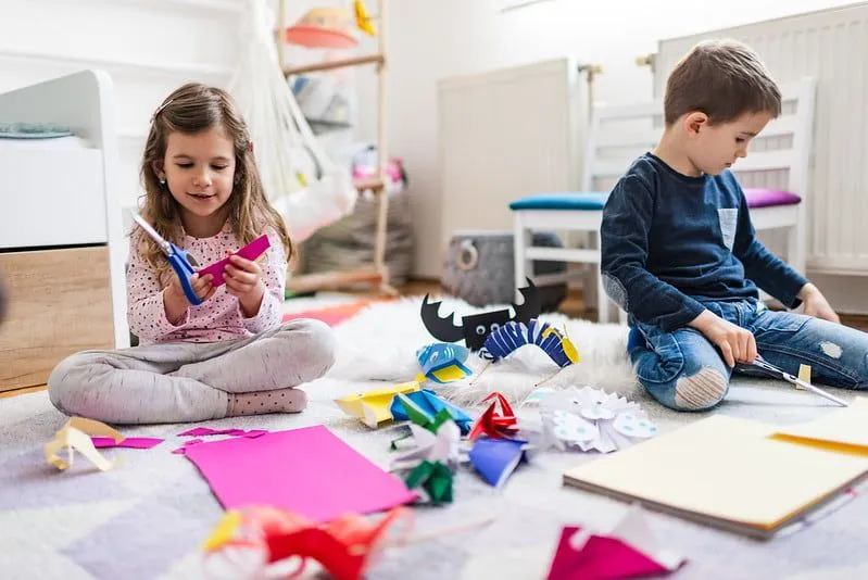 Two children sitting on the bedroom floor making paper crafts.