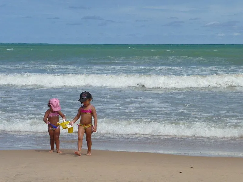 Two young children walking away from the sea up the sand carrying watering cans