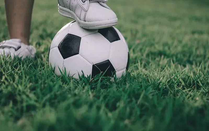 A child's foot on top of a football in the middle of a grassy field.