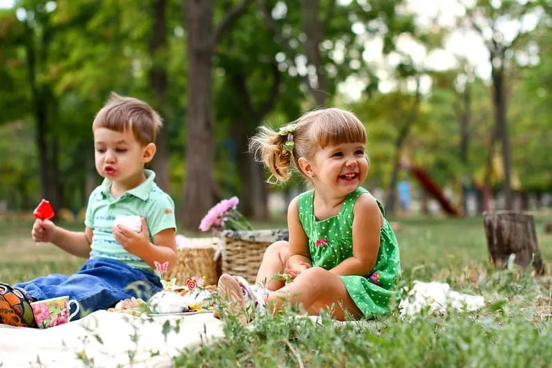 Little boy and girl sat on a rug on the grass outdoors enjoying a picnic.