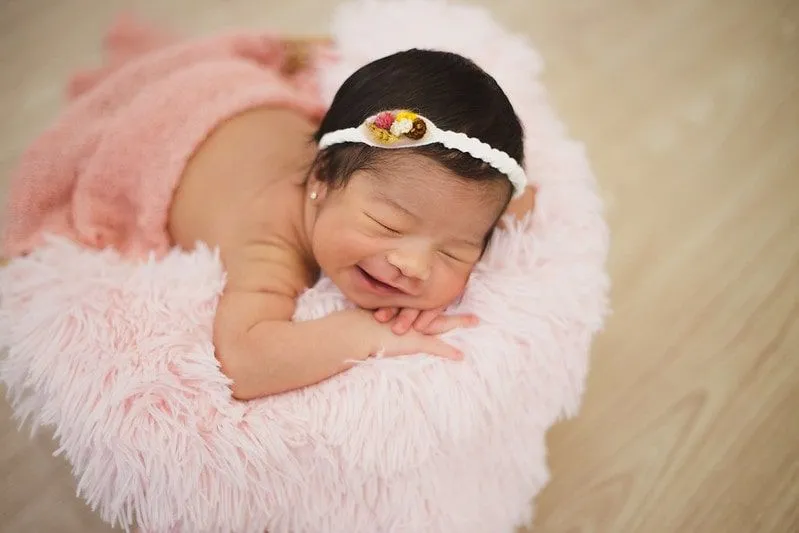 Baby girl wearing white woolly headband smiling lying on a pink blanket.