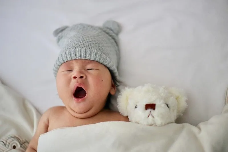 Baby boy wearing a woolly hat lying in bed next to a teddy, yawning.