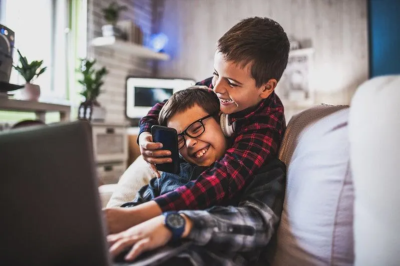 Two boys laughing while using gadgets on the sofa.