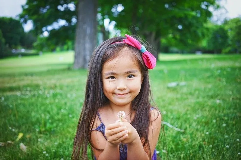 Little girl wearing a bow in her hair smiling and holding a flower.