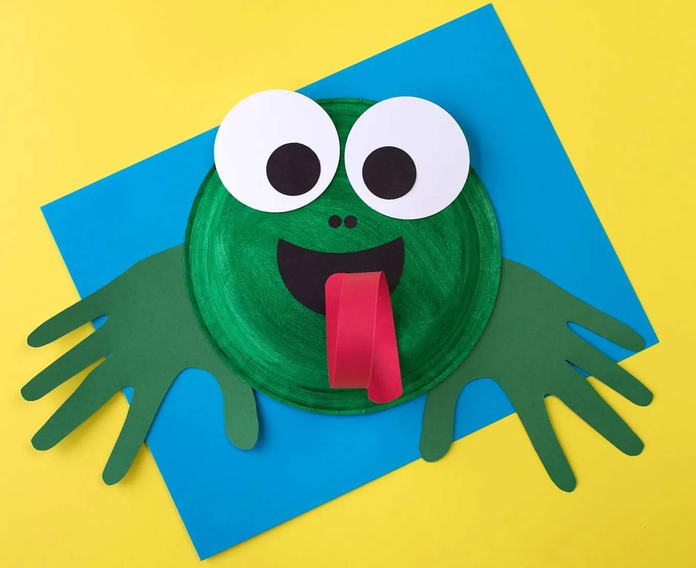Paper plate frog face with big googly eyes and its tongue sticking out.