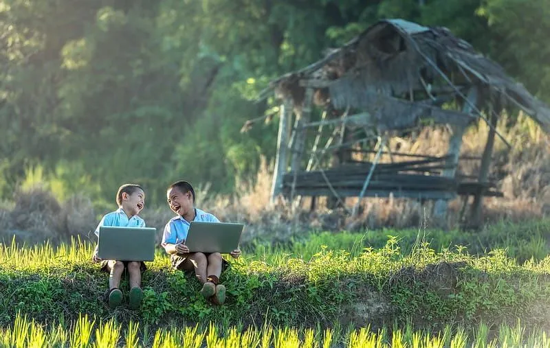 Two young boys sat outside with laptops looking at each other laughing.