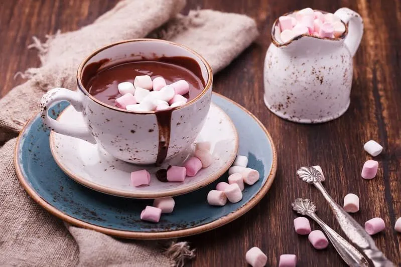 Cup of thick hot chocolate with marshmallows and a small jug filled with mini marshmallows on a wooden table.