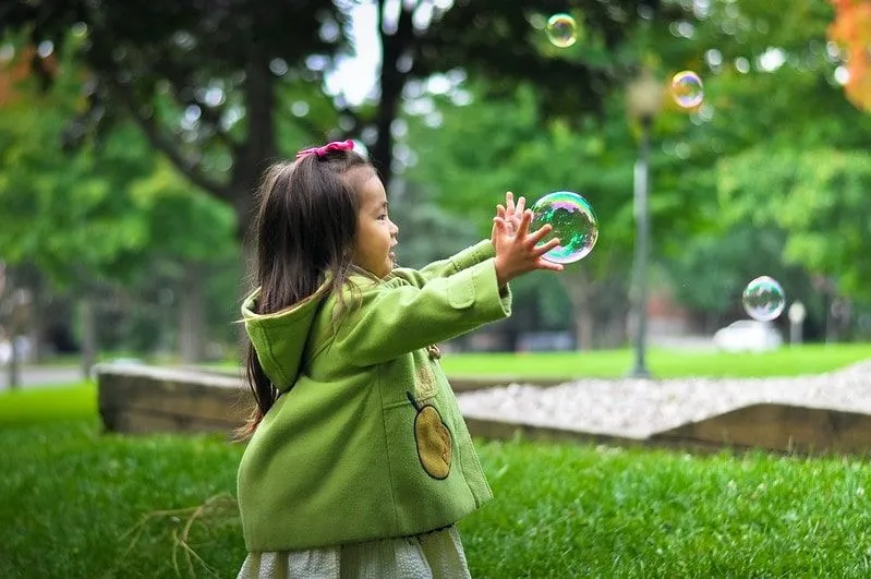 Little girl wearing a green jacket in the park reaching out to catch a bubble.
