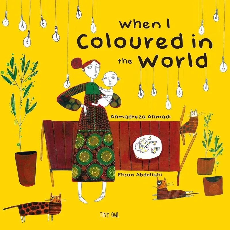 Cover of When I Coloured In The World: a woman in patterned green and brown clothing is holding a baby at her hip, both of which have no colour on their bodies. They are in a yellow room with brown furniture and three brown cats.