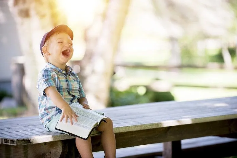 Little boy sat on top of a picnic bench with a book on his lap laughing.
