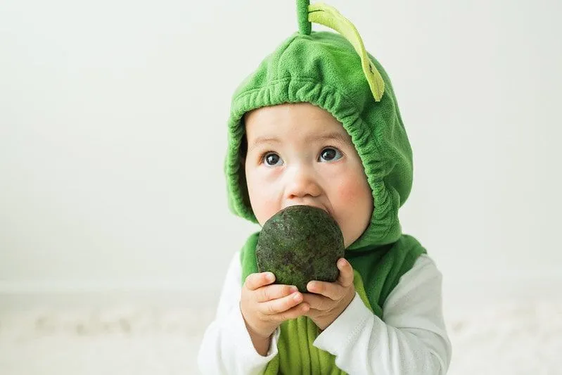 Baby wearing an avocado costume tries to eat a whole avocado.