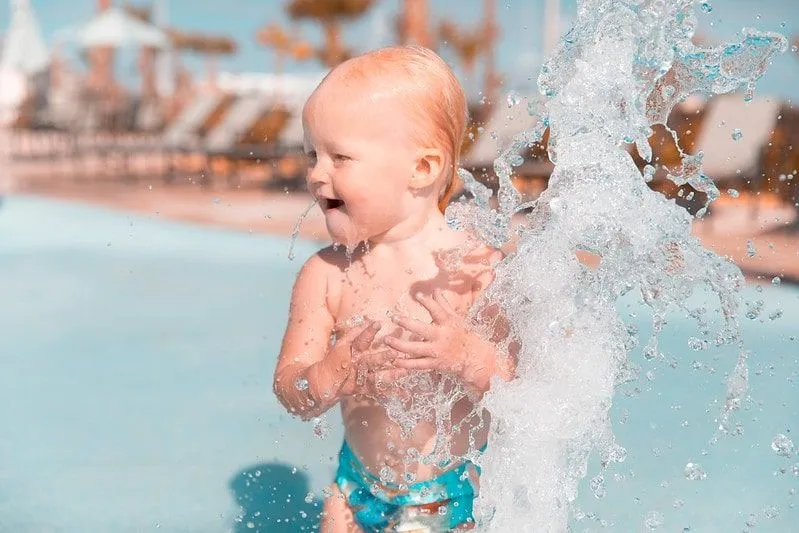 Toddler getting splashed in a water fountain at a water park laughing.