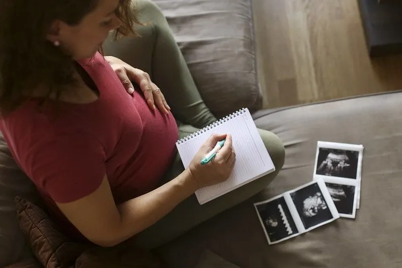 Pregnant mum sat on sofa with ultrasound photos writing in her baby scrapbook.