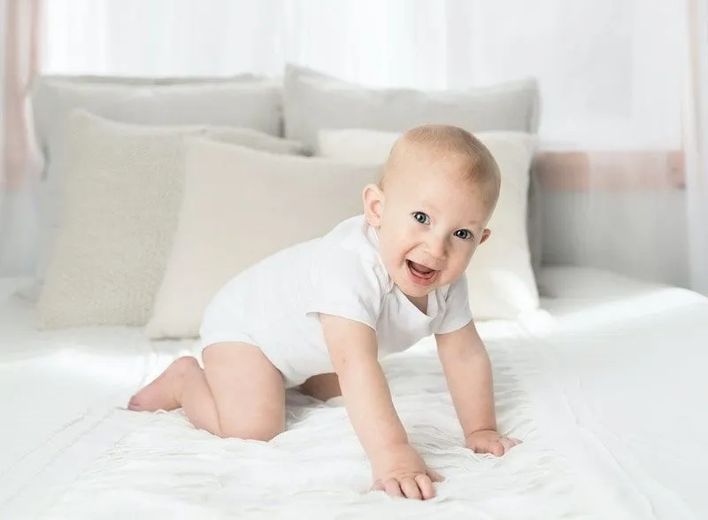 Baby wearing white crawling on the bed.