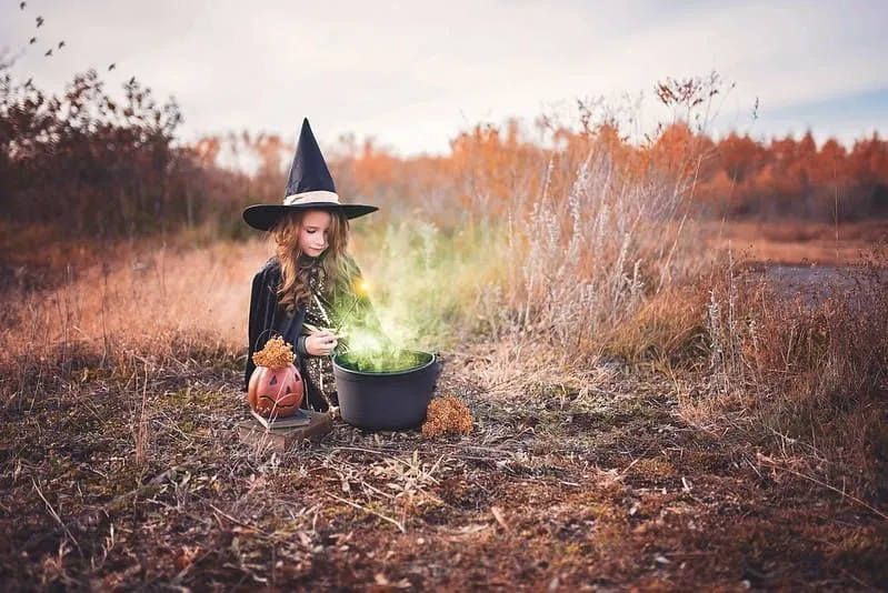 Little girl dressed as a witch sat on the ground in a field making a green potion in a cauldron.