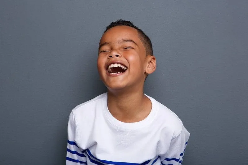 Young boy standing in front of a grey wall laughing at witch jokes.