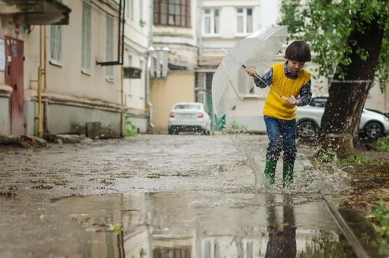 Young boy holding an umbrella splashes around in a puddle on the street in his wellies.