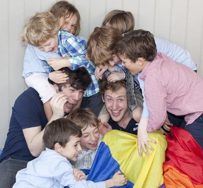 Lots of kids pile on top of two guys who are holding a parachute.