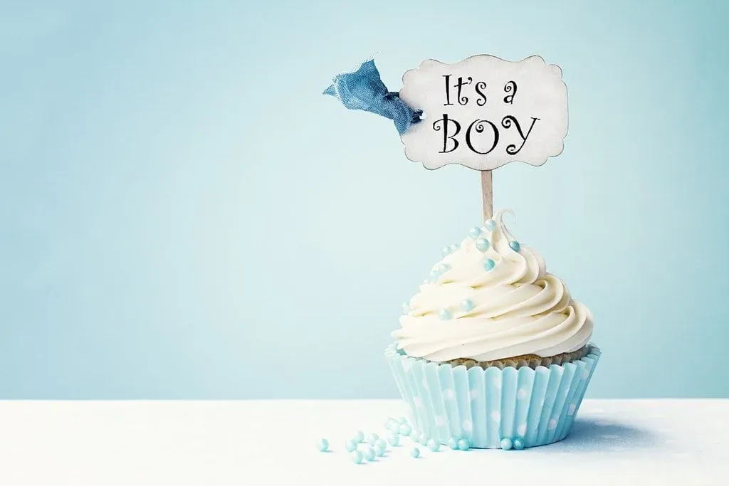 A cupcake in a blue cupcake case with blue sprinkles and a decoration saying 'It's a boy' on it. 