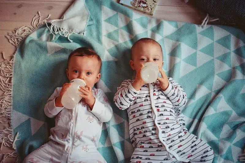 Two babies lying on a blanket on the floor drinking from their milk bottles.