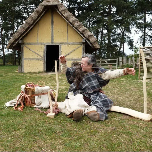 Woman in Anglo-Saxon village dressed as an Anglo-Saxon sat on the ground weaving.