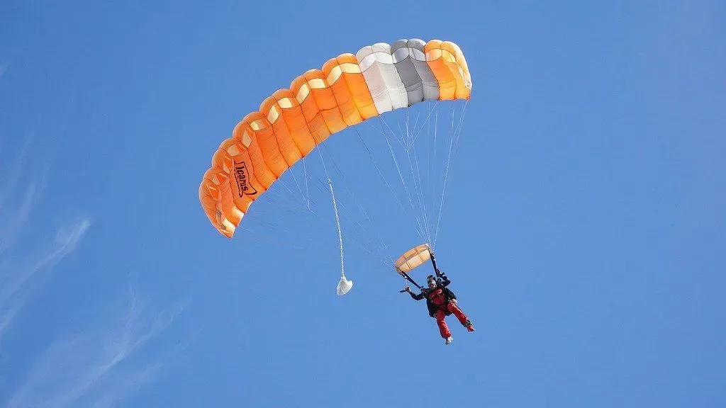 Man skydiving with an orange parachute is being pulled back down to the ground by gravity.
