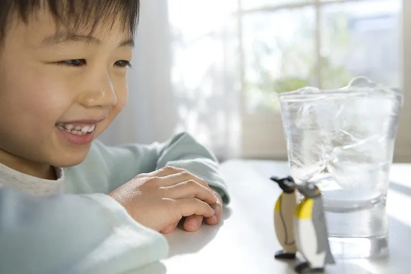 Boy sat at a table smiling at two toy penguins standing next to a glass of ice.
