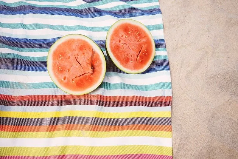 Two halves of a watermelon on a striped towel on the sandy beach.