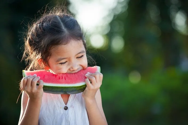 Girl biting into a wedge of watermelon, standing in the garden.