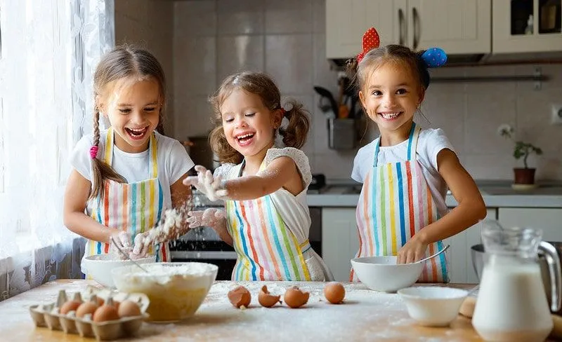 Three little girls in the kitchen smiling and laughing as they make pancakes.