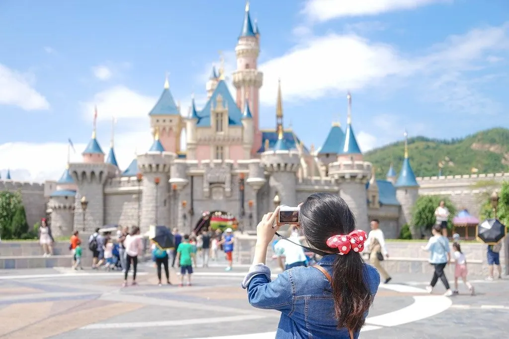 Girl wearing a Minnie Mouse scrunchie standing in front of the Disney castle taking a photo of it.