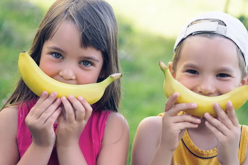 Little girl and boy standing outside holding up a banana to their face as a smile.
