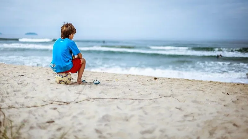 boy sat on football with flipflops in the sand