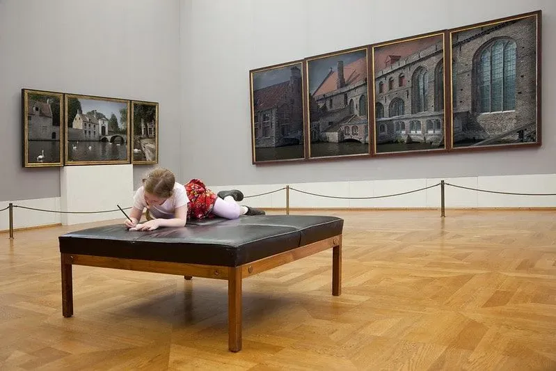 Little girl lying on a seat in a museum drawing in her notebook.
