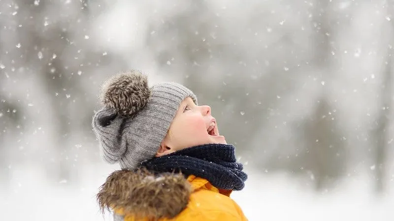 Little boy wrapped up wearing a grey woolly hat outside laughing as the snow falls.