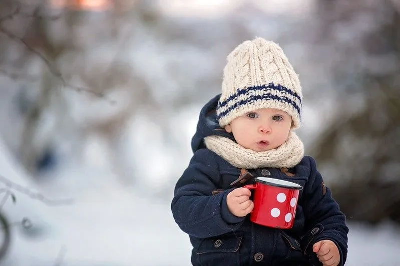 Baby boy wrapped up wearing a woolly hat, sat outside in the snow holding a mug.